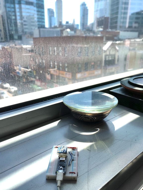 Figure 2. Nano 33 IoT and light sensor on a breadboard, positioned on a windowsill, overlooking a skyline of buildings in downtown Brooklyn. Sunlight streams in through the window, and the sensor is in partial shadow from the window frame.