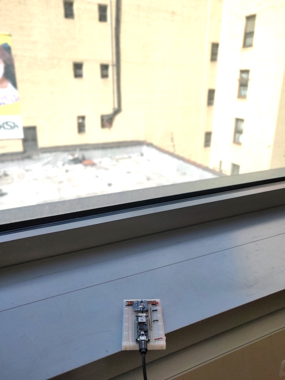 Figure 3. Nano 33 IoT and light sensor on a breadboard, positioned on a windowsill, overlooking a sunlit brick wall. the wall is painted a pale yellow.